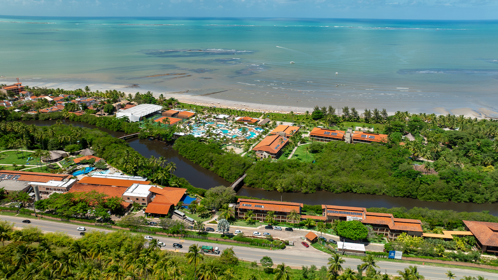 Imagem aérea do Salinas Maragogi mostrando o complexo de piscinas, dos quartos, pontos de consumo e ao fundo a praia. Área verde cercando todo o resort e o rio na parte inferior da imagem, é o rio que corta o resort.