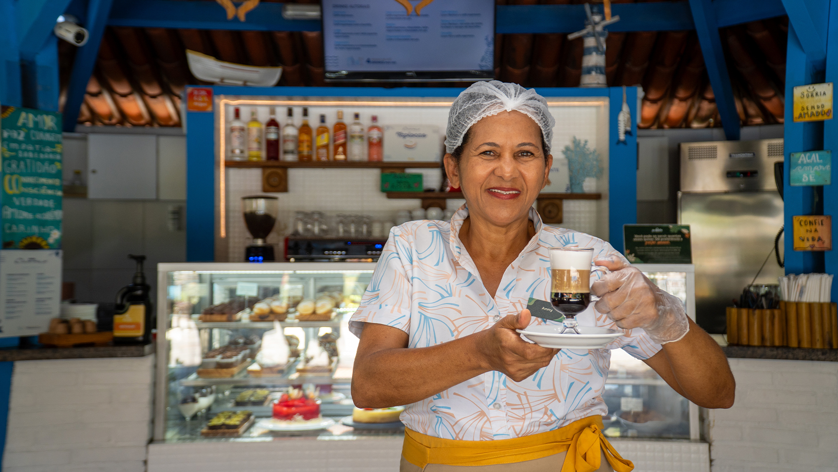Pessoa colaboradora do Salinas Maragogi servindo café especial no Café Canoas com vitrine de sobremesas ao fundo.