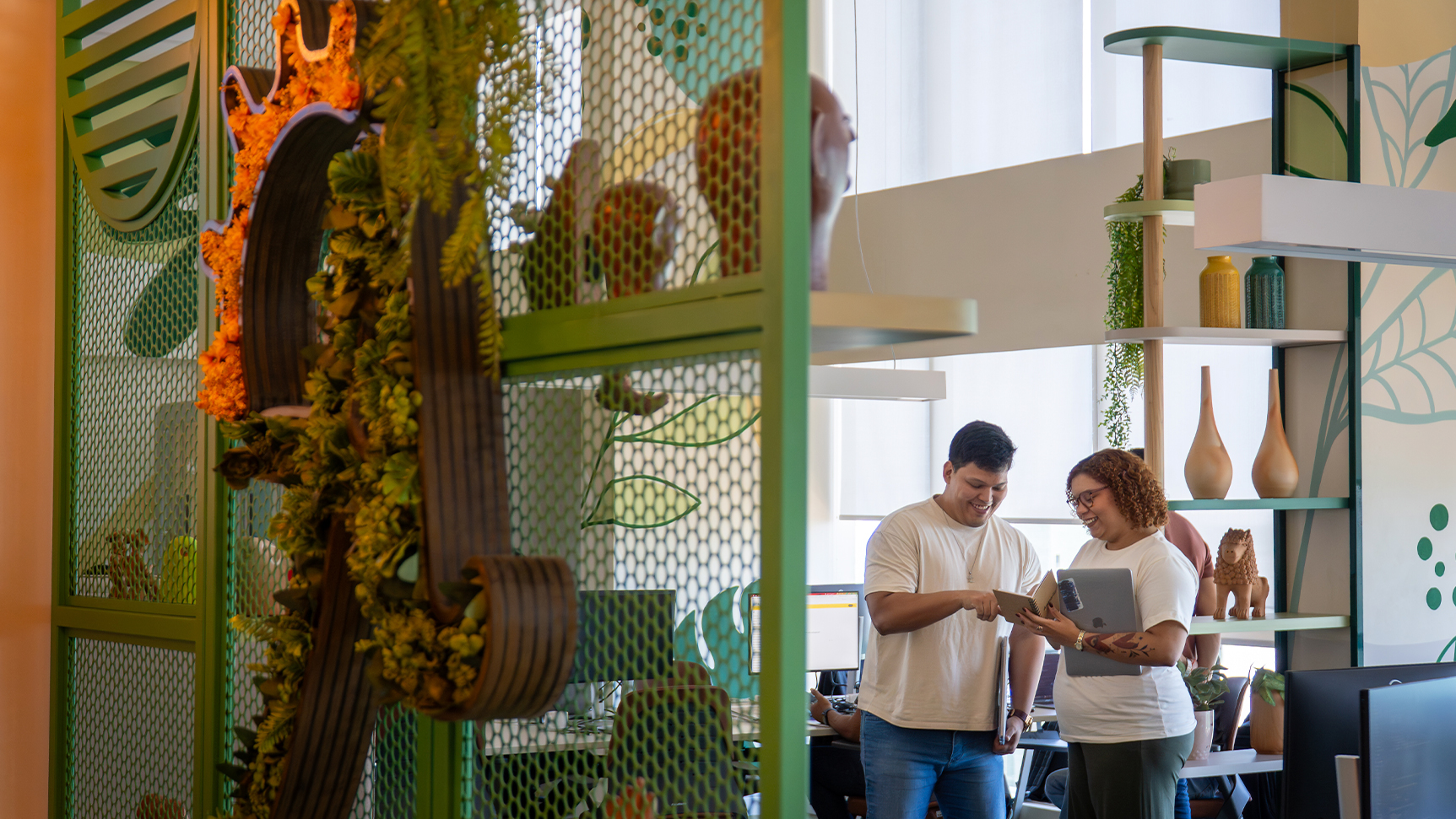 Colaboradores da Amarante sorriem enquanto conversam em um escritório moderno. Em primeiro plano, uma divisória verde com plantas e elementos orgânicos, ao fundo, um ambiente iluminado com decoração colorida mais verde.