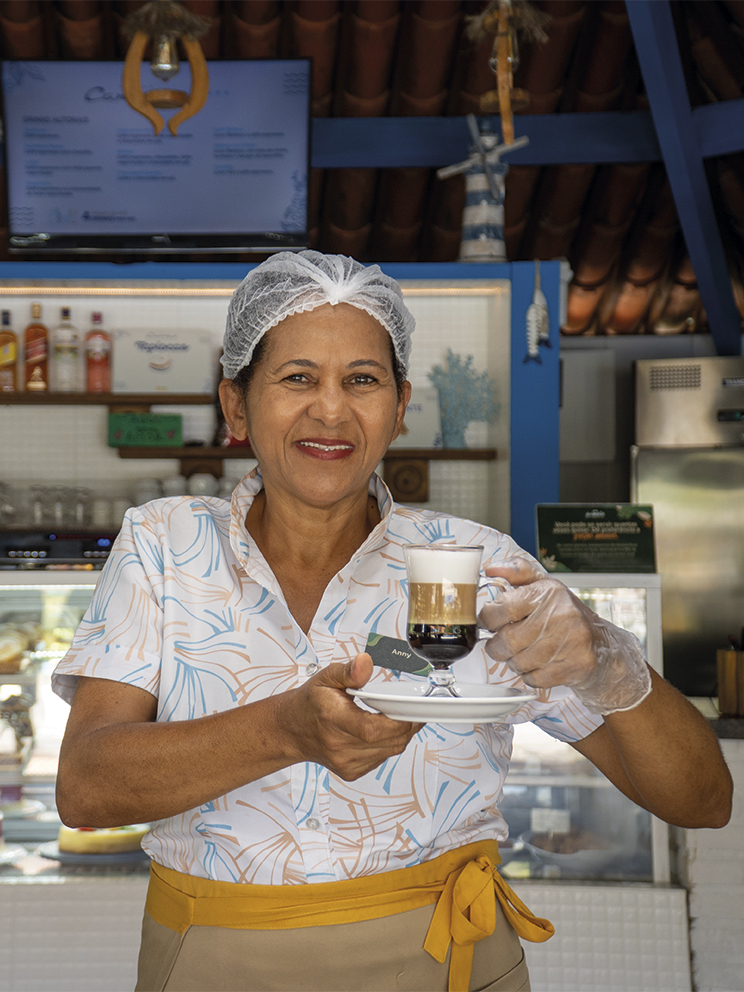Pessoa colaboradora do Salinas Maragogi servindo café especial no Café Canoas com vitrine de sobremesas ao fundo.