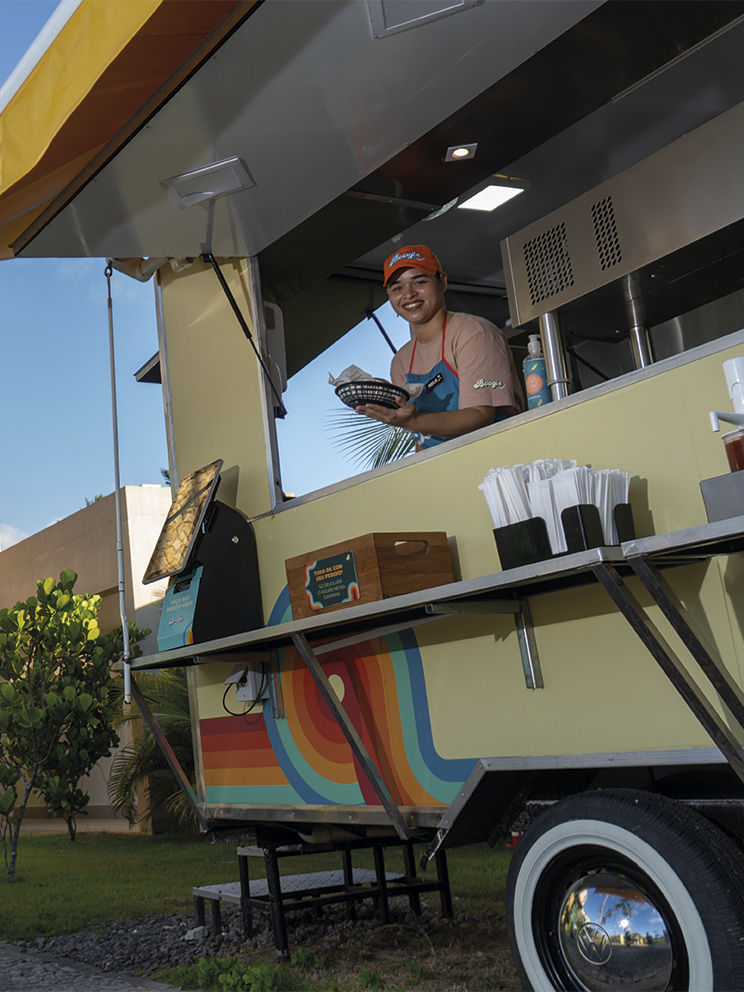 Colaboradora sorridente atende em um food truck colorido no Japaratinga Lounge Resort, entregando um lanche. Ao fundo, uma parede de pedras claras exibe o logotipo do Japaratinga Lounge Resort sob a luz do sol e sombras de palmeiras.