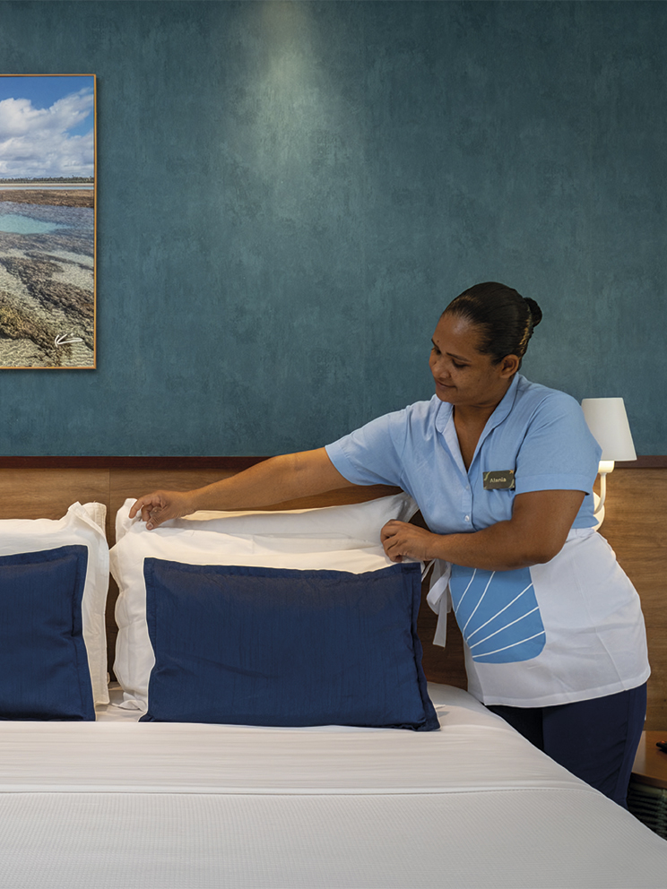 Colaboradora da governança do Salinas Maceió, usando uniforme azul e branco, arruma cuidadosamente os travesseiros de uma cama de casal em um quarto decorado com tons de azul e madeira.