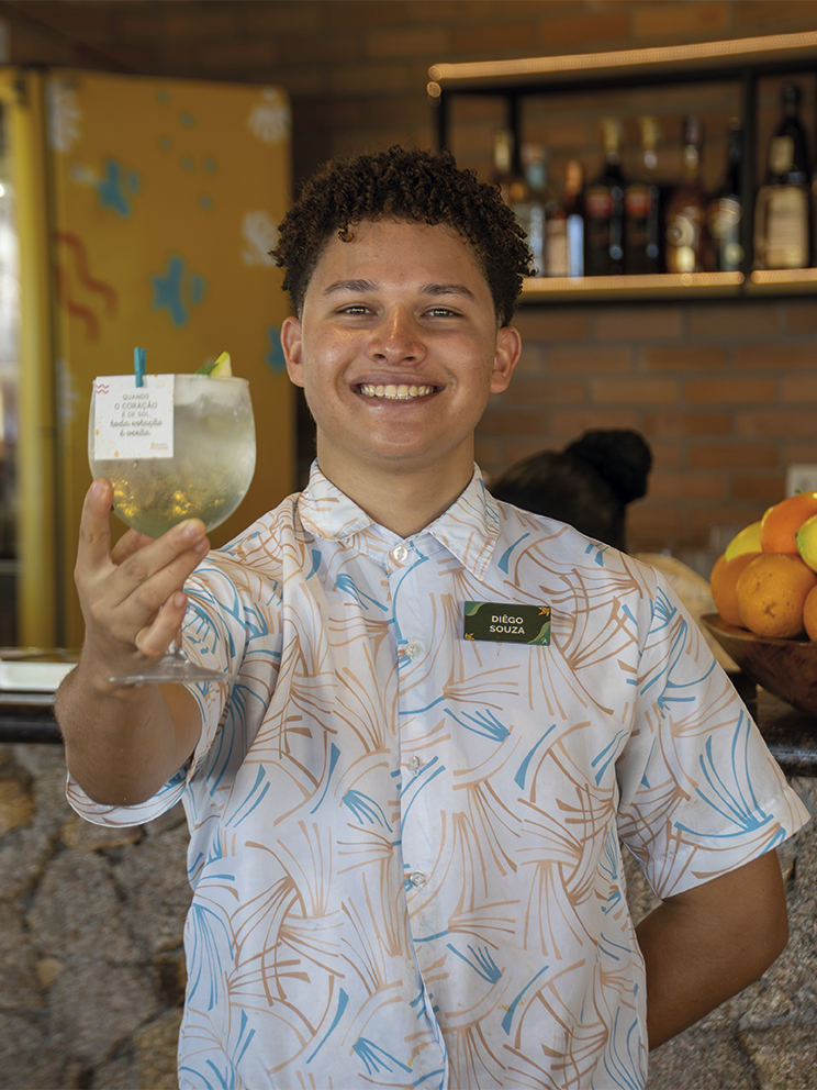 O colaborador do Salinas Maragogi, sorri segurando uma taça de drink refrescante em frente a um bar de pedras. Ele veste uma camisa estampada e ao fundo há uma fruteira tropical colorida.