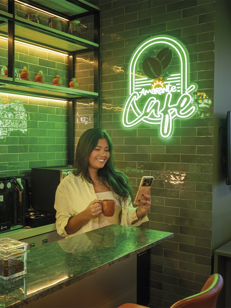 Mulher sorridente segura uma xícara de café e olha pro celular em um balcão moderno. O ambiente possui azulejos verdes, prateleiras de madeira com plantas e um letreiro neon escrito Amarante Café.