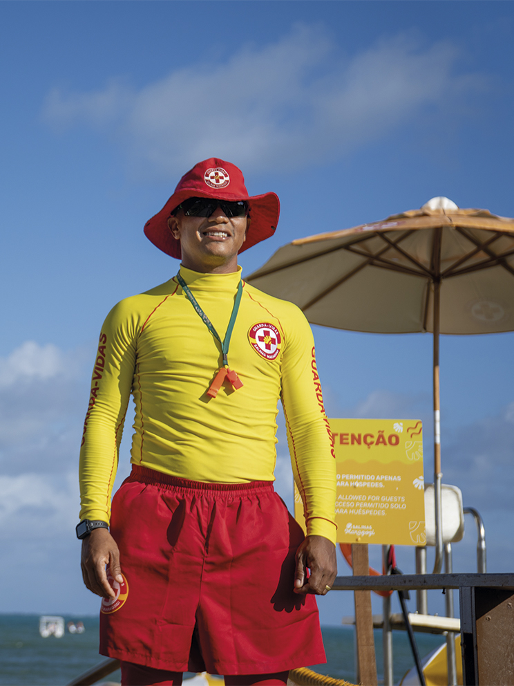 Salva-vidas do Salinas Maragogi sorri sob o sol, vestindo uniforme amarelo e vermelho com chapéu protetor. Ao fundo, o mar azul, céu com nuvens e guarda-sóis amarelos de praia.