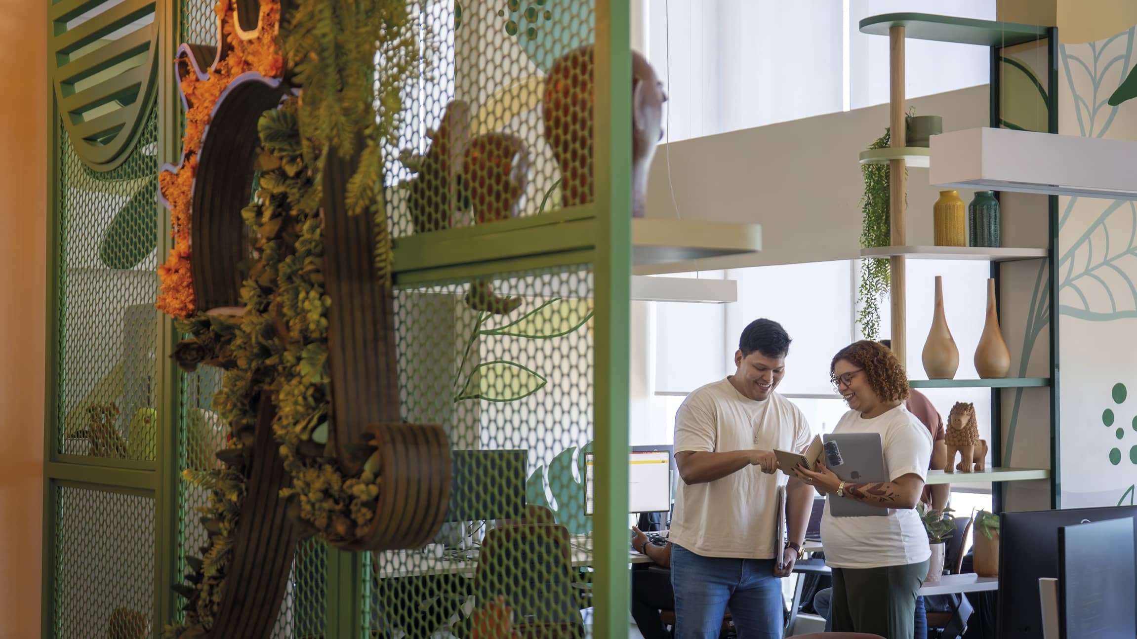 Colaboradores da Amarante sorriem enquanto conversam em um escritório moderno. Em primeiro plano, uma divisória verde com plantas e elementos orgânicos, ao fundo, um ambiente iluminado com decoração colorida mais verde.