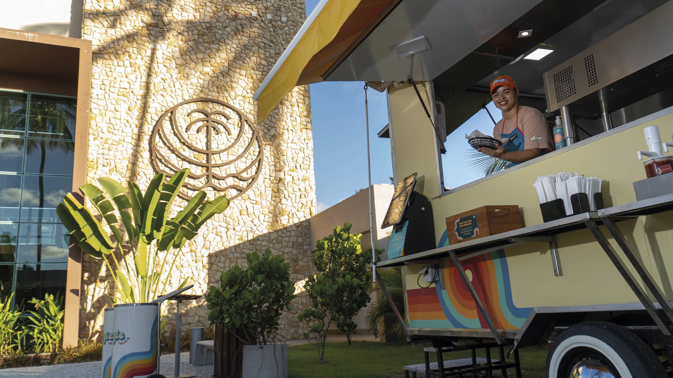 Colaboradora sorridente atende em um food truck colorido no Japaratinga Lounge Resort, entregando um lanche. Ao fundo, uma parede de pedras claras exibe o logotipo do Japaratinga Lounge Resort sob a luz do sol e sombras de palmeiras.