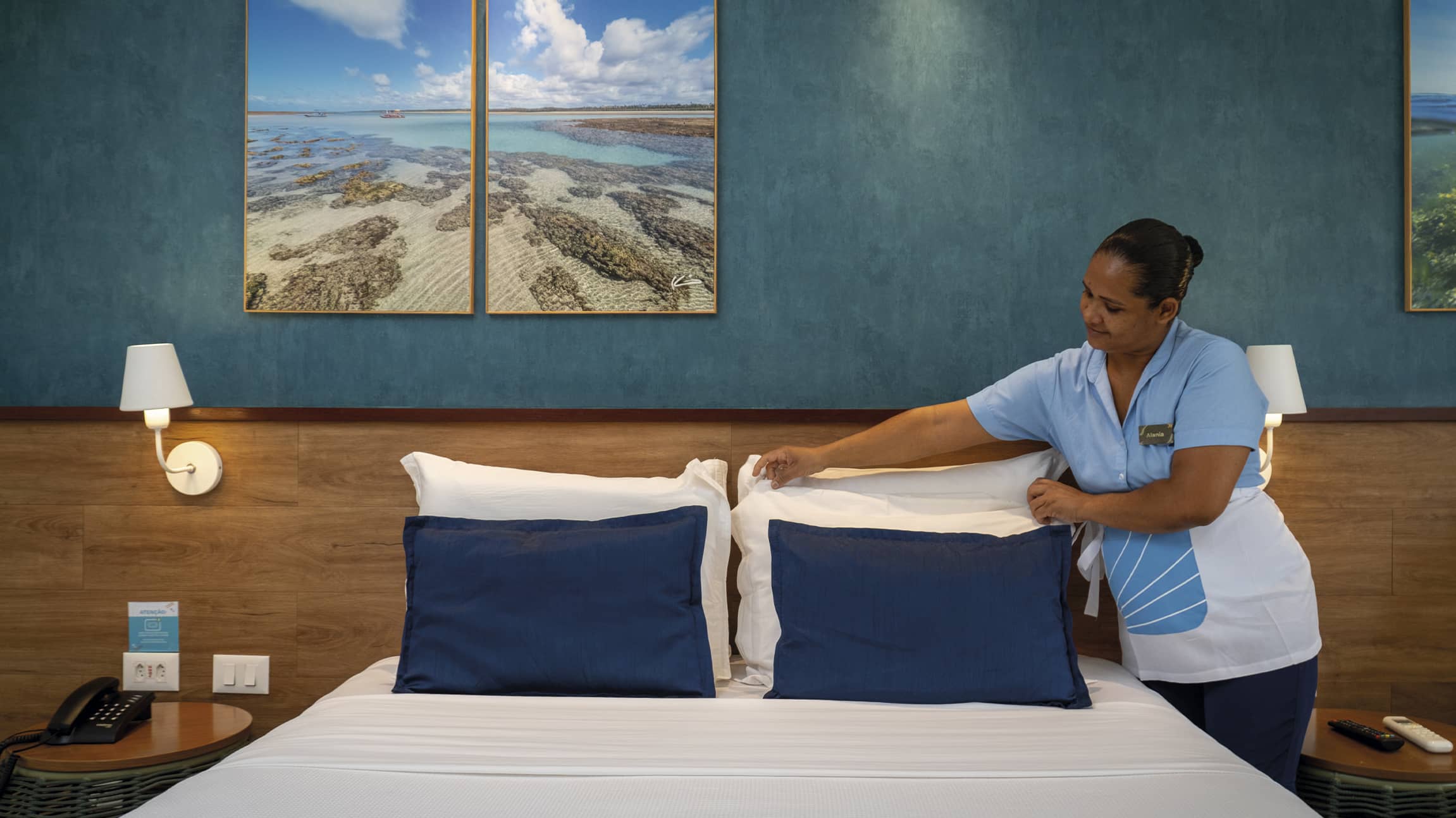 Colaboradora da governança do Salinas Maceió, usando uniforme azul e branco, arruma cuidadosamente os travesseiros de uma cama de casal em um quarto decorado com tons de azul e madeira.