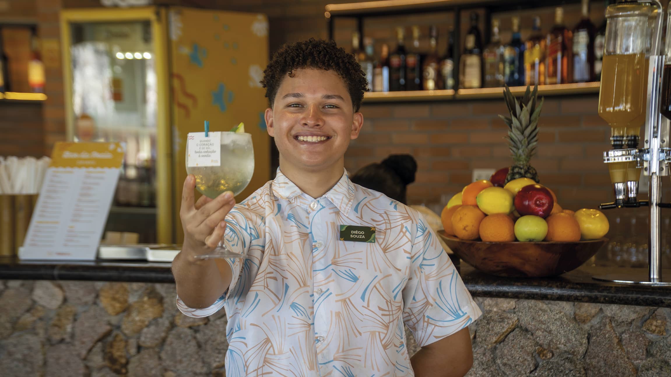 O colaborador do Salinas Maragogi, sorri segurando uma taça de drink refrescante em frente a um bar de pedras. Ele veste uma camisa estampada e ao fundo há uma fruteira tropical colorida.