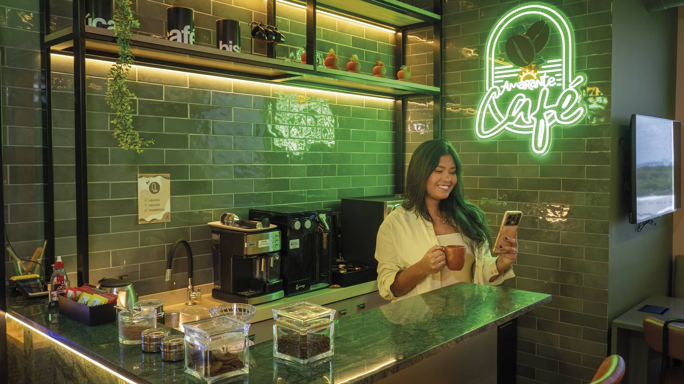 Mulher sorridente segura uma xícara de café e olha pro celular em um balcão moderno. O ambiente possui azulejos verdes, prateleiras de madeira com plantas e um letreiro neon escrito Amarante Café.