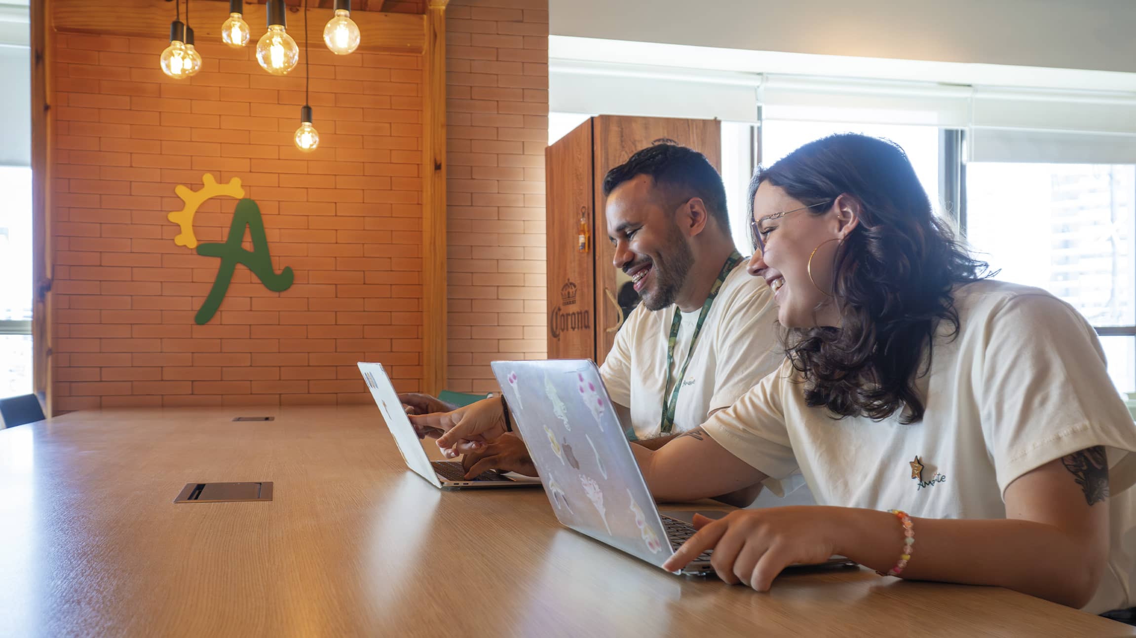 Dois colaboradores da Amarante sorriem enquanto trabalham juntos em notebooks em uma mesa de madeira. Ao fundo, parede de tijolinhos com o logotipo A estilizado e luminárias pendentes modernas.