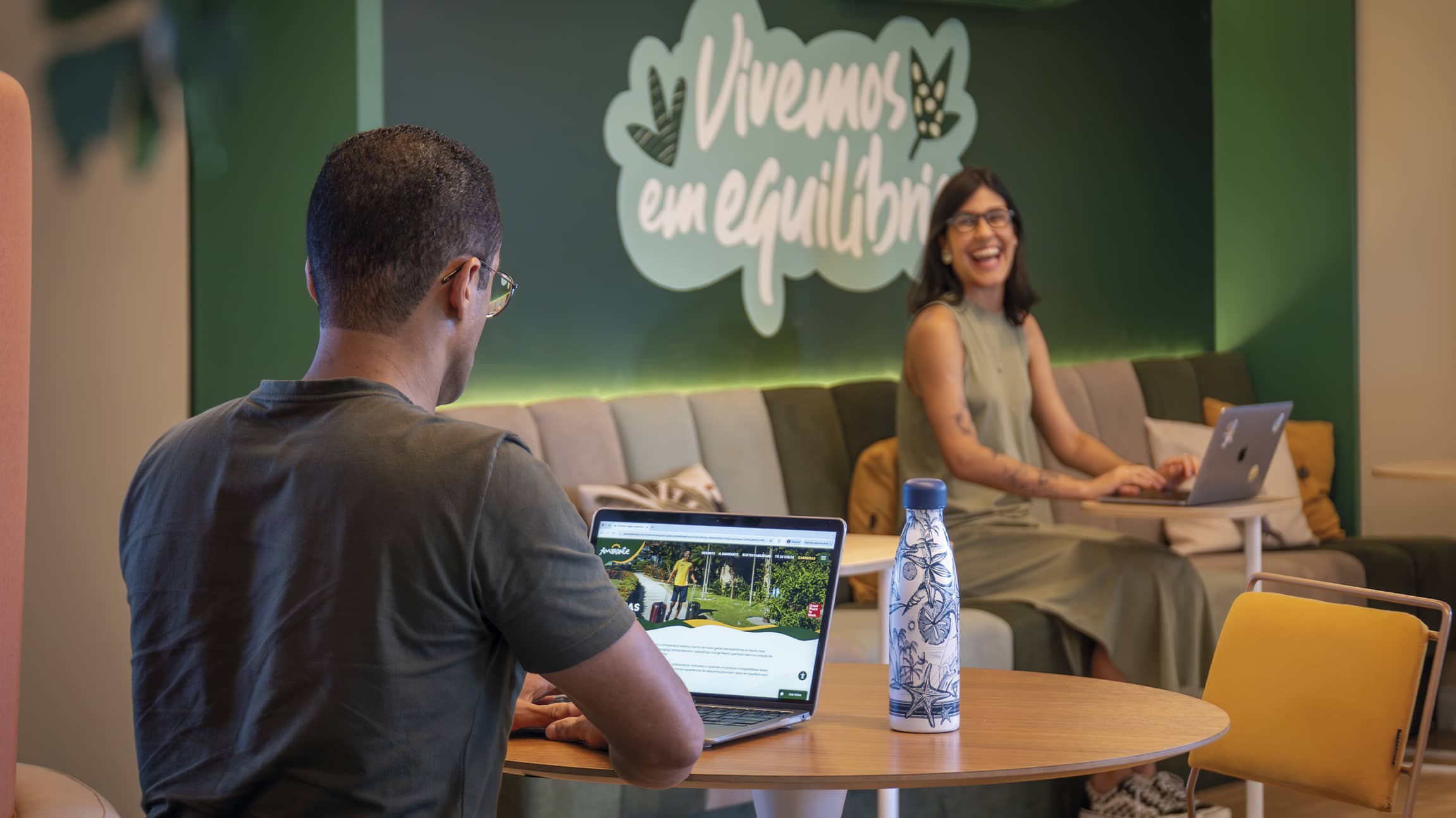 Ambiente de coworking com parede verde onde se lê Vivemos em equilíbrio. Um colaborador em primeiro plano e uma colaboradora ao fundo sorriem enquanto trabalham em um espaço acolhedor.