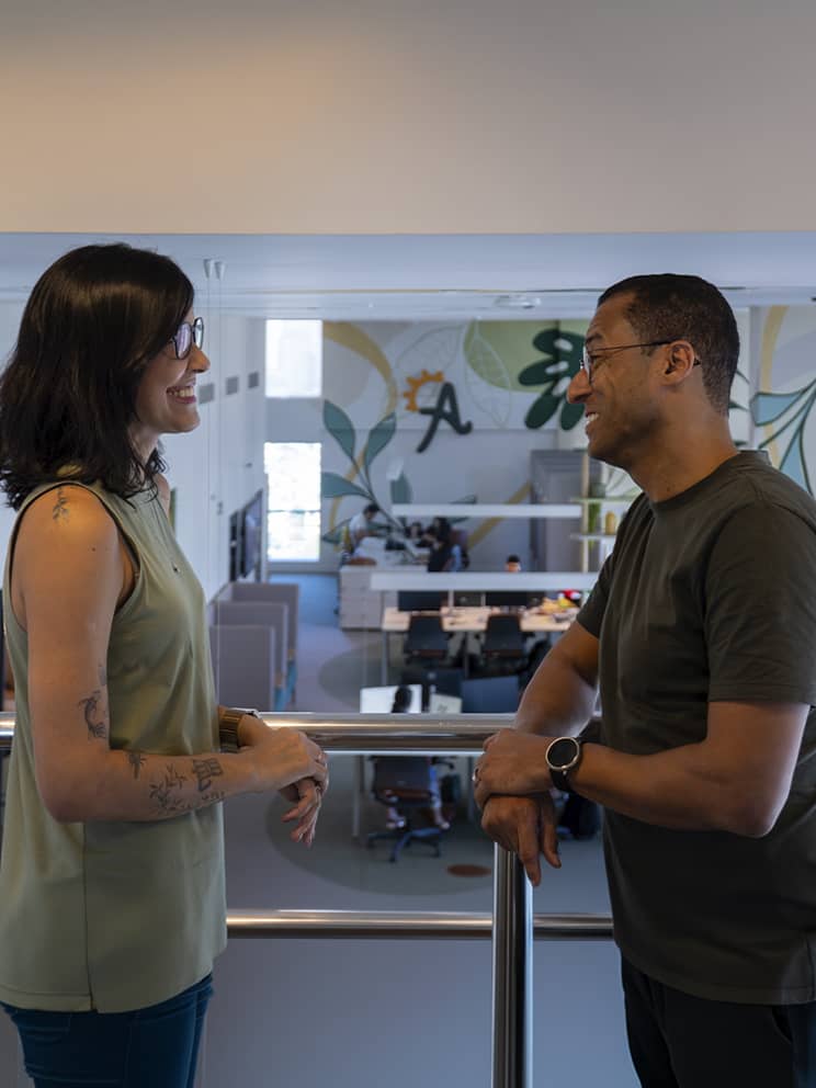 Pessoas colaboradoras da Amarante interagindo no escritório de Recife. Decoração com plantas e um ambiente leve e acolhedor. Uma grande que separa a recepção do escritório tem a logo da Amarante.