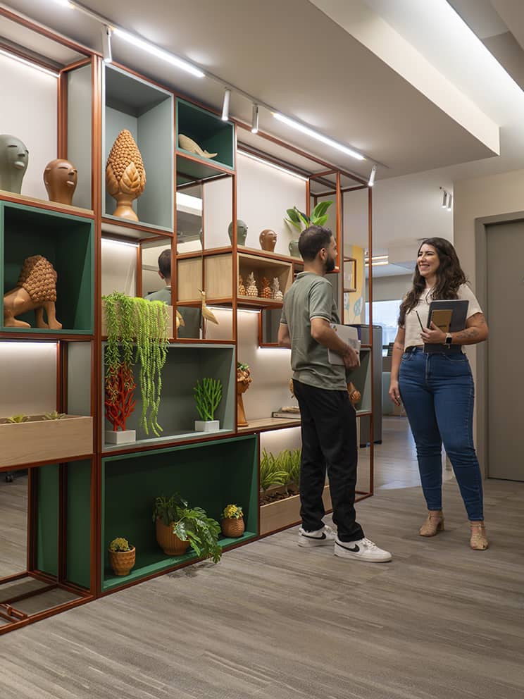 Corredor do escritório corporativo da Amarante com duas colaboradoras andando e conversando, ao lado direito um letreiro da Amarante e uma baia de trabalho com funcionários e computadores, e ao lado esquerdo uma prateleira de decoração.