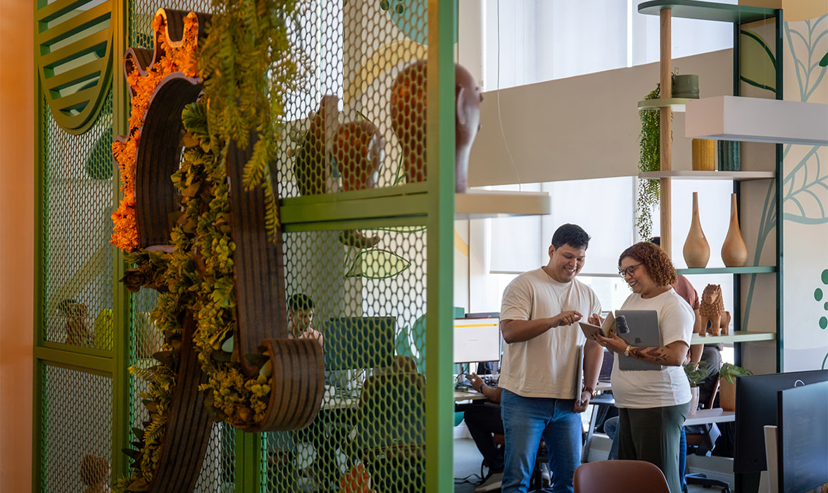 Colaboradores da Amarante sorriem enquanto conversam em um escritório moderno. Em primeiro plano, uma divisória verde com plantas e elementos orgânicos, ao fundo, um ambiente iluminado com decoração colorida mais verde.