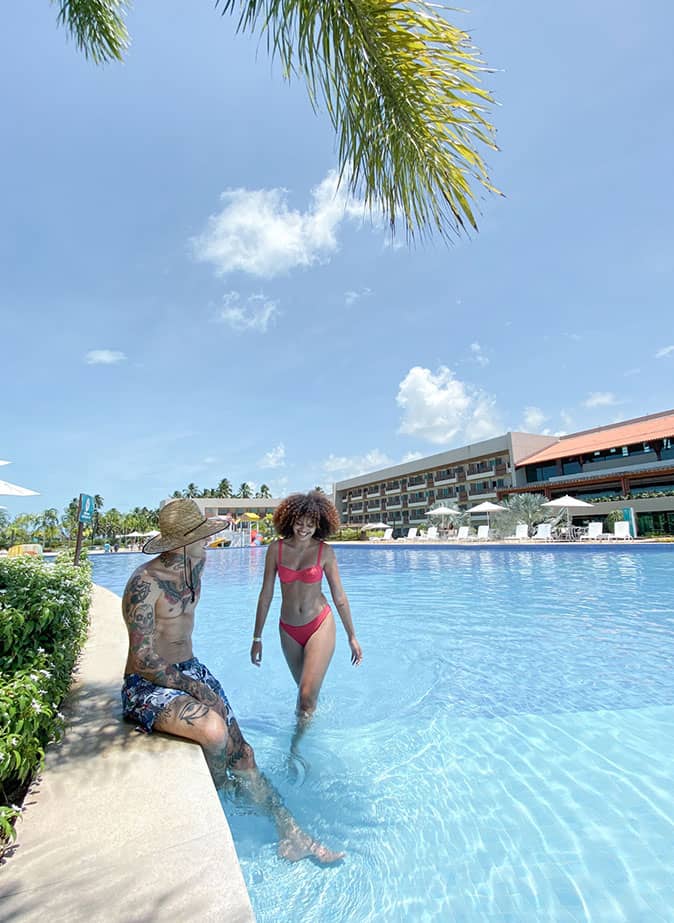 Um casal de homem e mulher está andando na beira de uma das piscinas do Japaratinga Lounge Resort. Água de uma cor azul, assim como o céu quase sem nuvens nuvens.