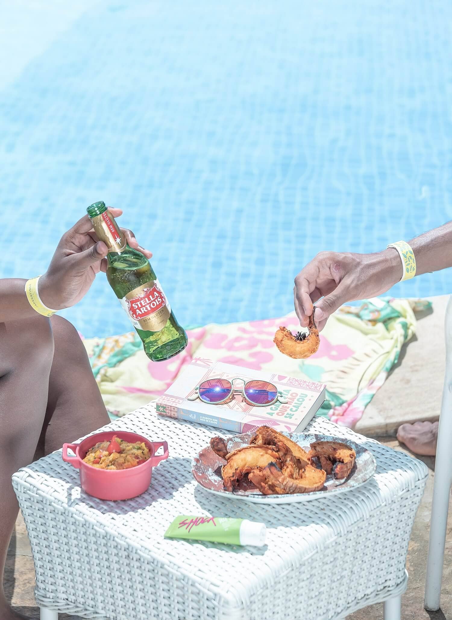 Na imagem é possível ver parte de duas pessoas desfrutando do all inclusive premium do Japaratinga Lounge Resort, resort no Nordeste do Brasil. Há petiscos de camarão e uma cerveja long neck, ao fundo vê-se uma piscina.