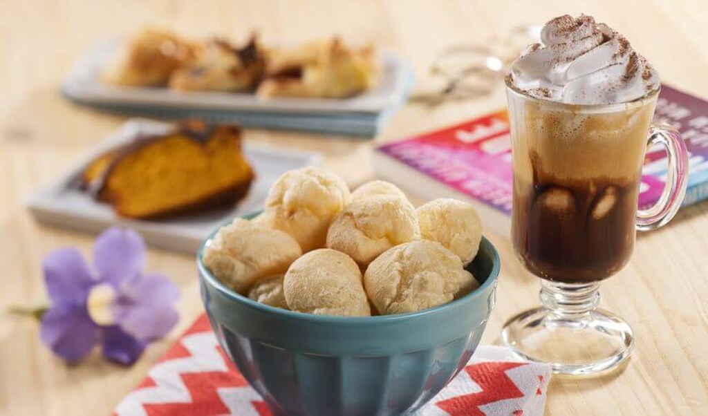 Pão de queijo e uma bebida quente com chantilly em uma mesa de café da manhã, com bolos caseiros ao fundo.