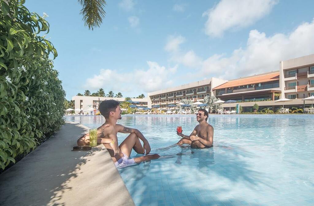 Dois homens relaxando e tomando drinks na piscina de um resort luxuoso sob céu claro e clima ensolarado.