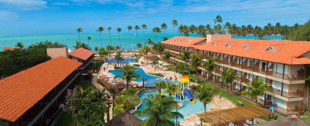Vista do Resort Salinas Maceió, com piscina e área de praia ao fundo, cercada por palmeiras e céu azul.
