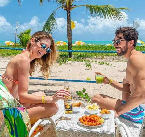 Casal feliz saboreando uma refeição à beira da praia, com o mar ao fundo e o sol brilhando.