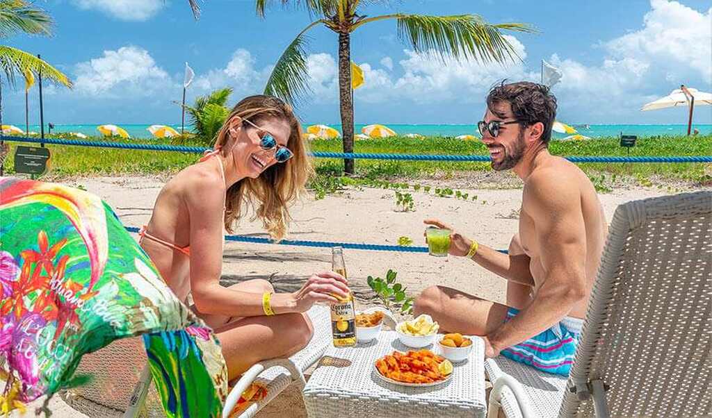 Casal feliz saboreando uma refeição à beira da praia, com o mar ao fundo e o sol brilhando.