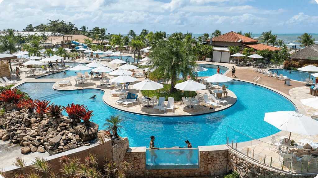 Vista panorâmica de um resort com piscina aquecida, rodeada por coqueiros, espreguiçadeiras e bar molhado em frente ao mar em Alagoas.