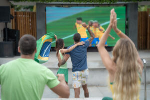 Quatro pessoas de costas, incluindo um casal abraçado com a bandeira do Brasil, torcem e comemoram em frente a um telão LED gigante que exibe um jogo de futebol (jogadores com uniformes amarelos e azuis) no Japaratinga Lounge Resort.