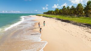 Família correndo na faixa de areia da praia do Gunga, em Maceió. Mar azul turquesae ampla faixa de coqueiros presentes nas zonas laterais da imagem.