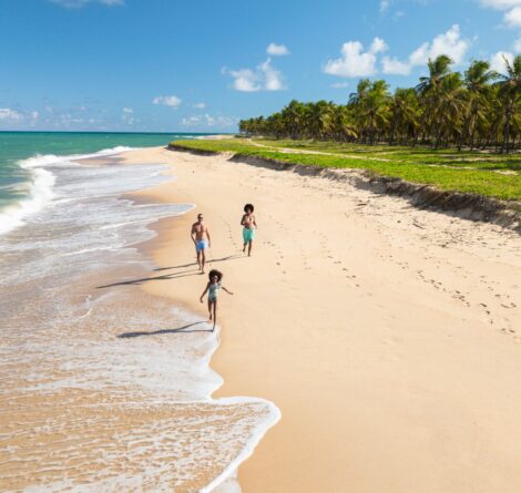 Família correndo na faixa de areia da praia do Gunga, em Maceió. Mar azul turquesae ampla faixa de coqueiros presentes nas zonas laterais da imagem.