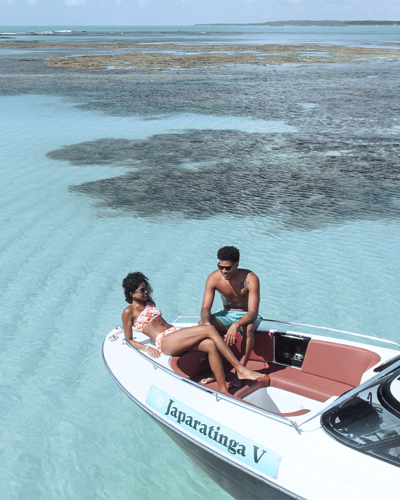 Passeio de barco pelas piscinas naturais de Japaratinga Casal relaxando a bordo de um barco nas piscinas naturais de Japaratinga, em Alagoas, com o mar em tons de azul turquesa.