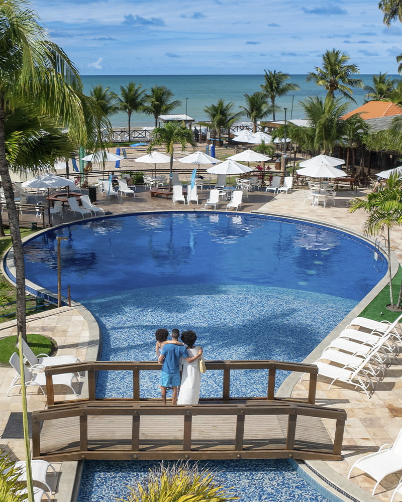 A vista que te espera no Salinas Maceió Resort All Inclusive Casal admirando a piscina do Salinas Maceió, com deck de madeira, espreguiçadeiras e o mar ao fundo.