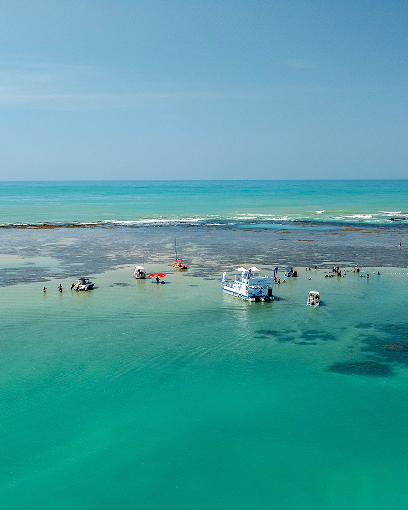 Praia de Pajuçara com águas cristalinas Vista aérea da Praia de Pajuçara com mar cristalino, recifes de corais visíveis e algumas embarcações atracadas nas piscinas naturais.
