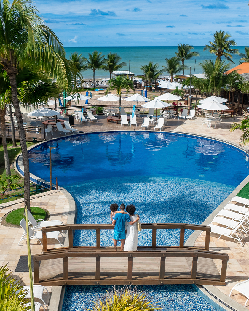 Piscinas do Salinas Maceió pra todas as idades Piscina redonda, com ponte de madeira passando por cima dela. Na ponta há uma família de pai, mãe e filha observando o horizonte do resort e praia do Salinas Maceió cercada por coqueiros e áreas de lazer.