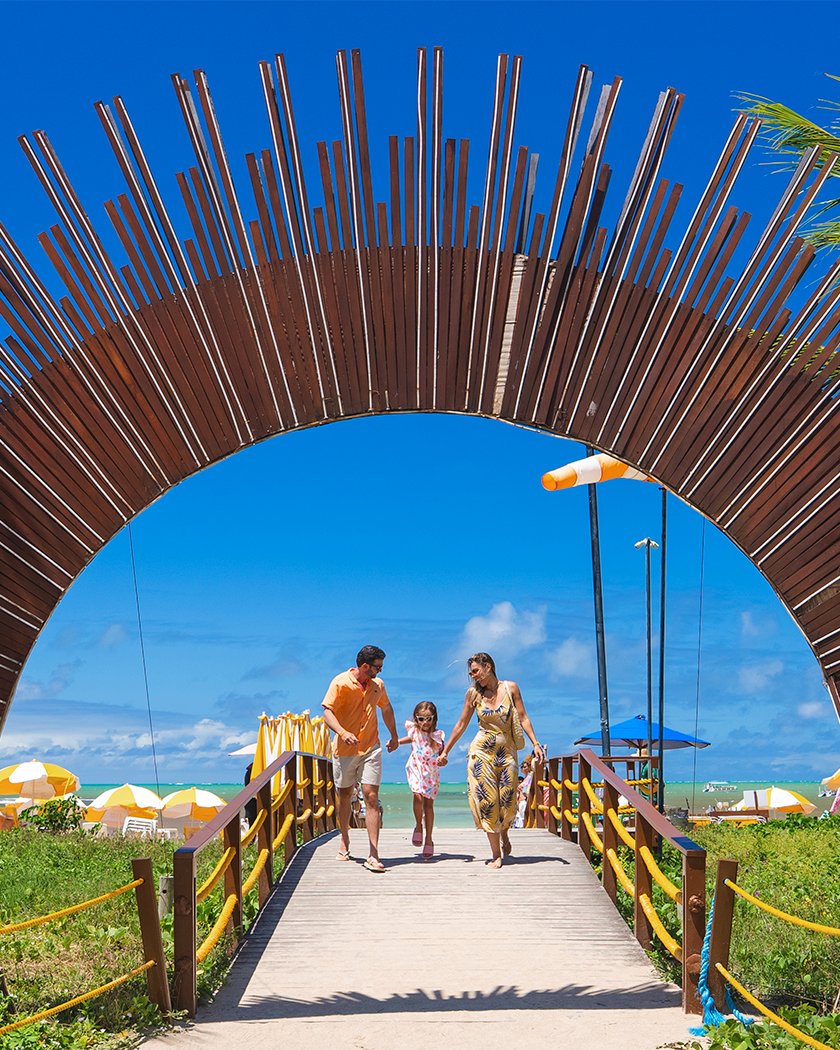 Onde as férias começam Portal do Salinas Maragogi cercado por coqueiros na beira do mar com céu azul.