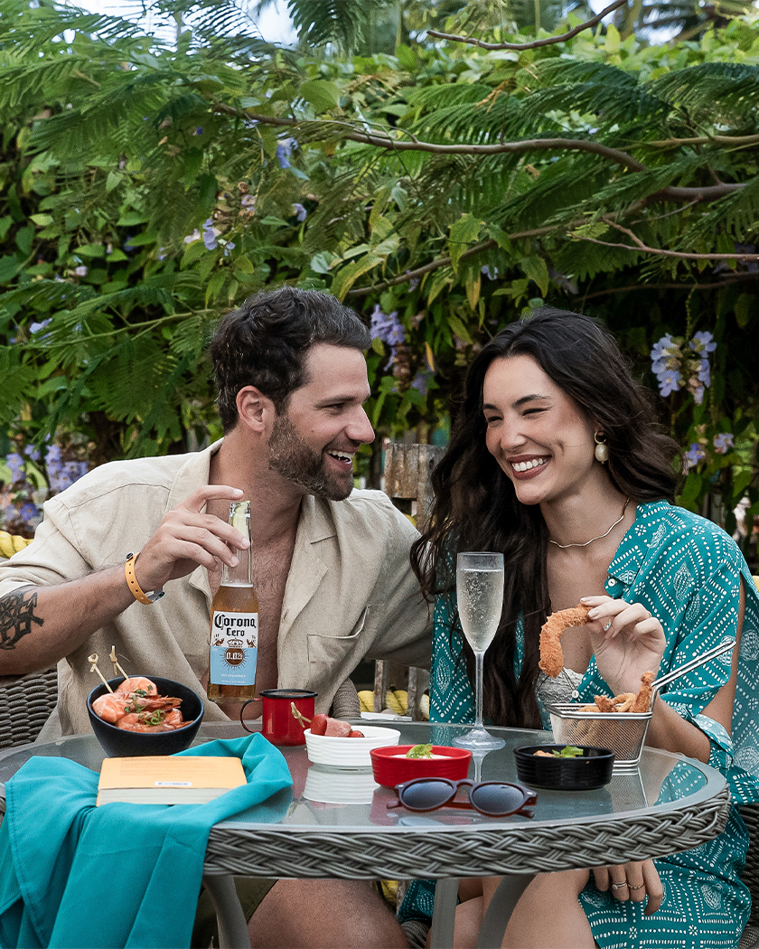 all inclusive premium 24h dia Casal no Vila da Praia em mesa com cerveja long neck Corona Extra ao lado de um taça de champanhe, com petiscos como camarão no espeto e camarão empanado, ao fundo muitas plantas com flores lilás.