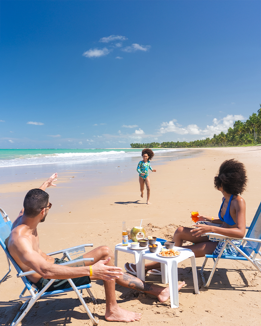 praia de ipioca alagoas Família relaxando em uma praia de areia clara. O casal está sentado em cadeiras de praia, com petiscos e bebidas em uma mesa. A filha corre e sorri na areia vinda da direção do mar. Um coqueiral e o mar azul-turquesa estão no fundo.