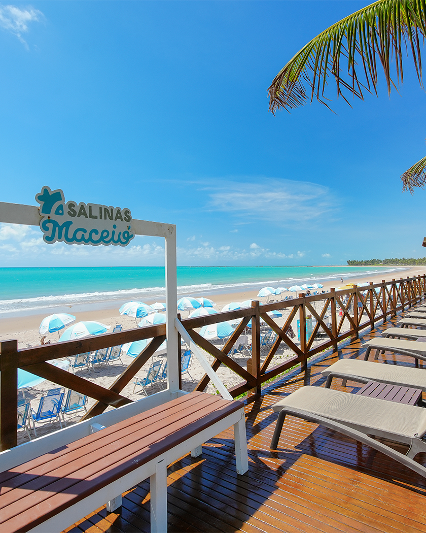 destinos paradisíacos ipioca Vista do deck de madeira do resort Salinas Maceió, com espreguiçadeiras beges, à beira do mar. A praia de Ipioca azul claro, e areia clara com guarda-sois listrados branco e azuis.
