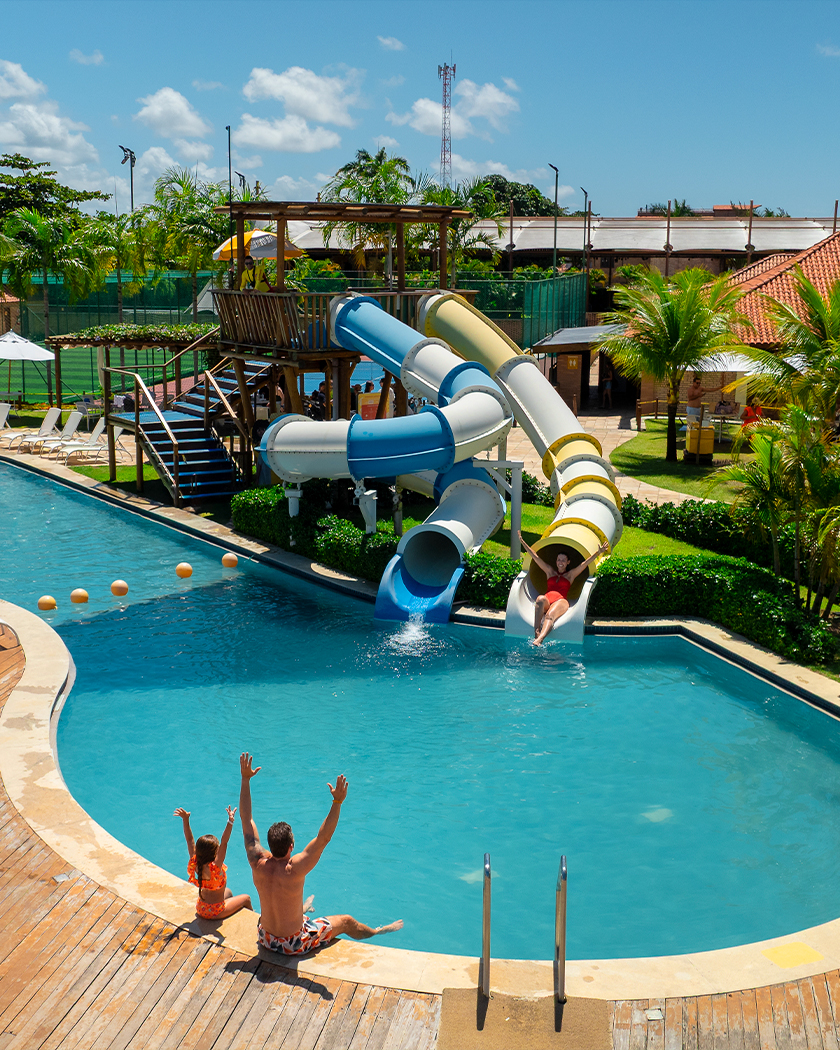 estrutura completa salinas maragogi Pai e filha sentados na beira da piscina do Salinas Maragogi. Na piscina tem estrutura de toboáguas infantis, e ao fundo estruturas do resort.