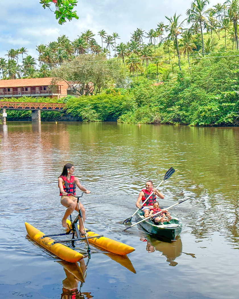 atividades lazer caiaque Rio que cruza o Salinas Maragogi com família fazendo atividade de caiaque e bike boat, cercado por manguezais, e ao fundo um pedaço da estrutura do resort.