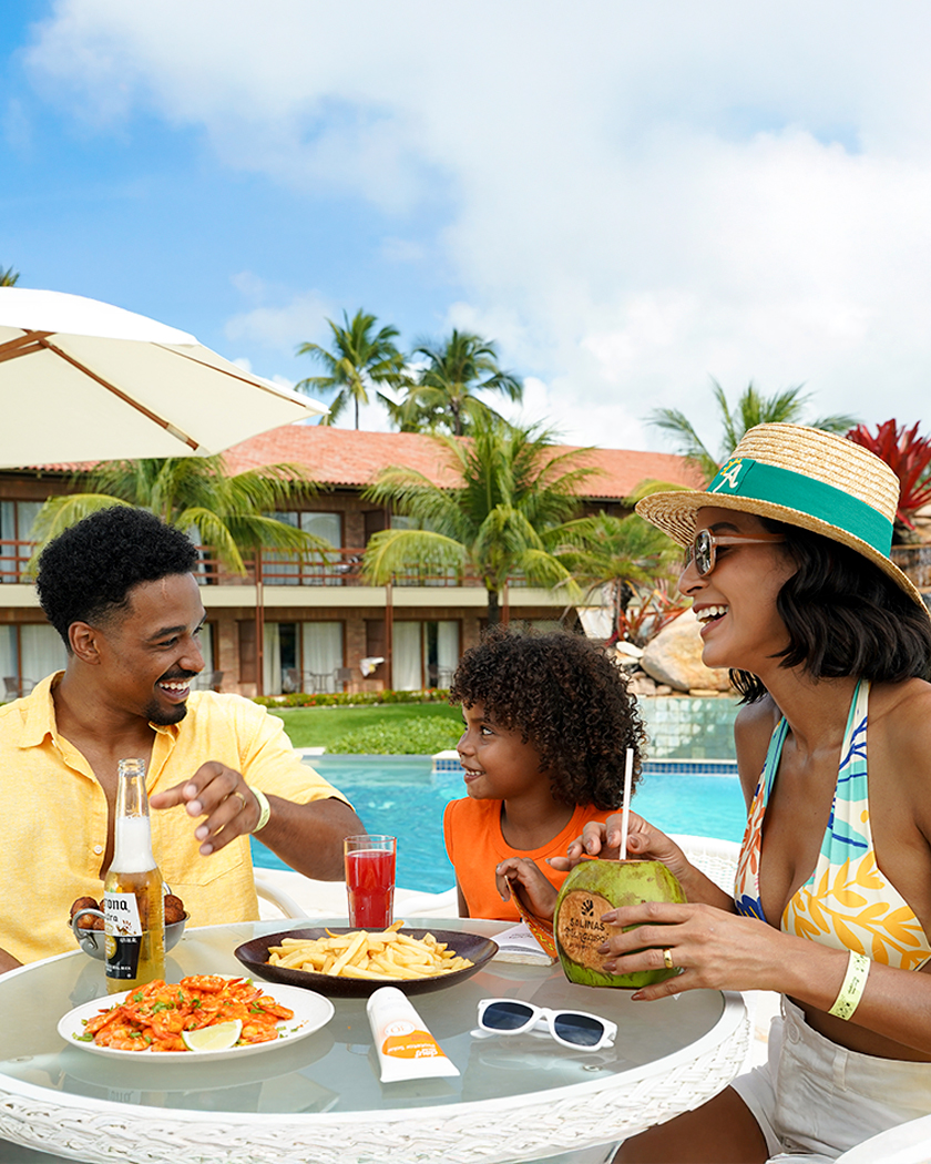 familia all inclusive Família sorrindo e petiscando à beira da piscina em um dia ensolarado. A mesa redonda está cheia de pratos com camarões e batatas fritas. O pai tem uma long neck Corona e a mãe está com um água de coco, e sentado com um suco vermelho. Ao fundo, coqueiros, a piscina e a estrutura do resort Salinas Maragogi.