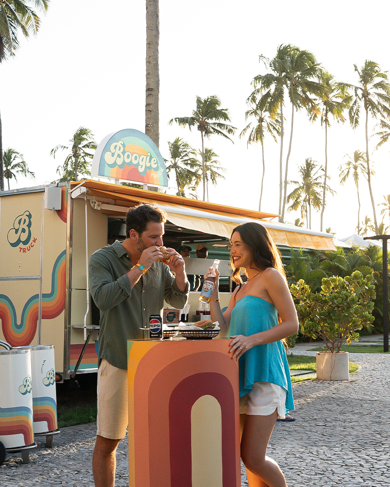 Aquele hambúrguer que não pode faltar nas férias Casal se divertindo no food truck Boogie do Japaratinga Lounge Resort, comendo e bebendo ao ar livre, rodeado por palmeiras.