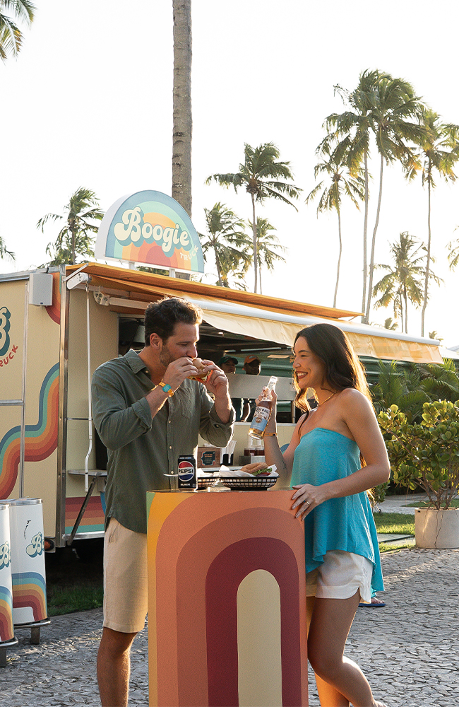 Aquele hambúrguer que não pode faltar nas férias Casal se divertindo no food truck Boogie do Japaratinga Lounge Resort, comendo e bebendo ao ar livre, rodeado por palmeiras.