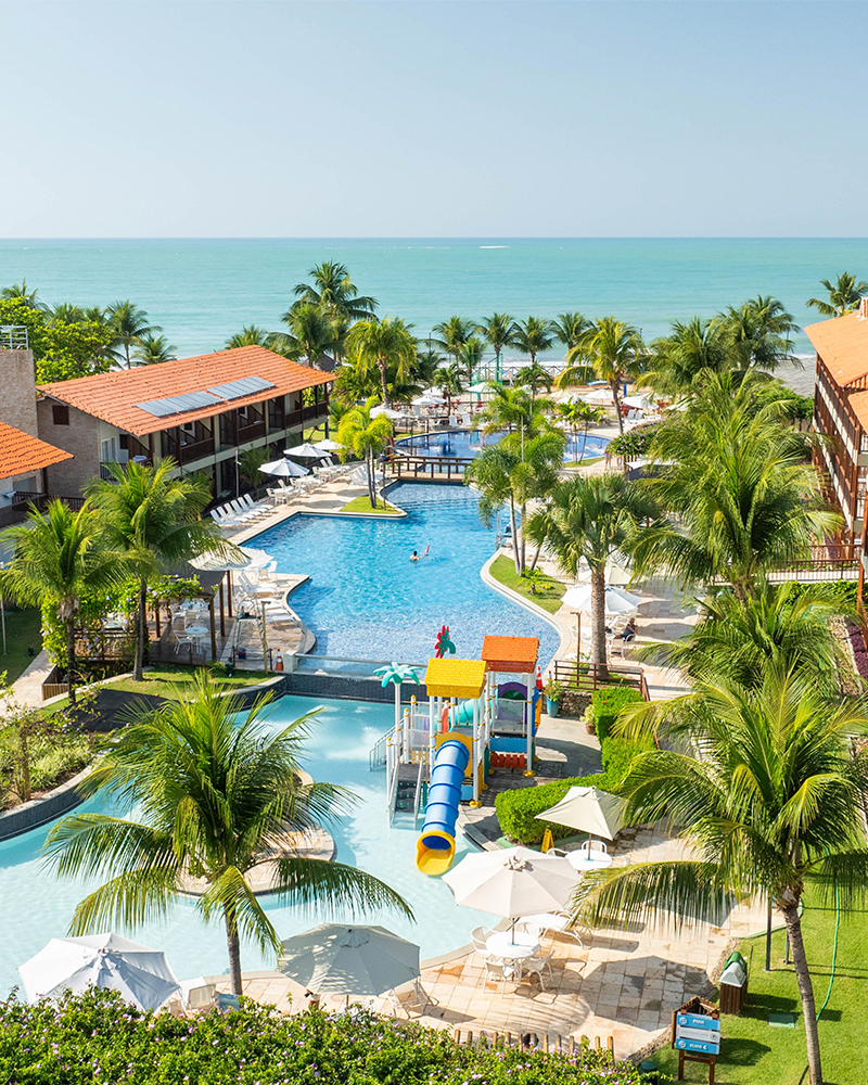 Diversão pra todos da família no Salinas Maceió Vista aérea de um resort Salinas Maceió pé na areia com prédios de telhados laranjas. A área de lazer inclui piscinas com decks, coqueiros e um parque aquático infantil com escorregadores coloridos. O mar está visível ao fundo.