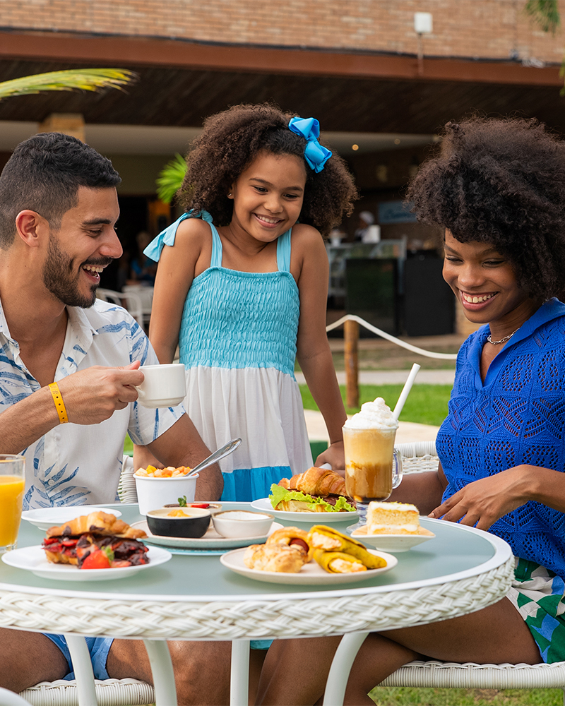 Tudo que o melhor All Inclusive pode oferecer Família sorrindo, sentada em mesas brancas no gramado do resort Salinas Maceió, desfrutando de um lanche com sanduíches, salgados, doces e bebidas. O pai segura uma caneca de café, a mãe uma bebida gelada e a filha observa.