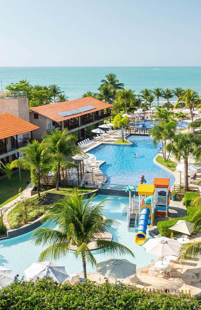 Diversão pra todos da família no Salinas Maceió Vista aérea de um resort Salinas Maceió pé na areia com prédios de telhados laranjas. A área de lazer inclui piscinas com decks, coqueiros e um parque aquático infantil com escorregadores coloridos. O mar está visível ao fundo.