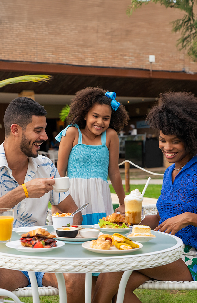 Tudo que o melhor All Inclusive pode oferecer Família sorrindo, sentada em mesas brancas no gramado do resort Salinas Maceió, desfrutando de um lanche com sanduíches, salgados, doces e bebidas. O pai segura uma caneca de café, a mãe uma bebida gelada e a filha observa.