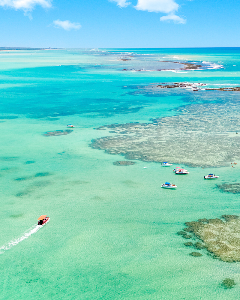 Piscinas naturais de Barra Grande que se transforma em cartão-postal Vista aérea das piscinas naturais de Barra Grande, com águas cristalinas e formações de corais, barcos ancorados e turistas desfrutando do mar em cenário paradisíaco.
