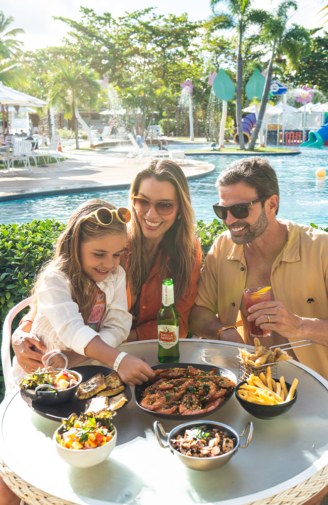 Aquele All Inclusive 24h que agrada desde a criançada até os papais. Família sorrindo e almoçando à beira da piscina em um dia ensolarado. A mesa redonda está cheia de pratos com camarões, batatas fritas e saladas. O pai segura um drink e a mãe está com a filha no colo e tem uma garrafa de cerveja na mesa. Ao fundo, coqueiros, a piscina e um playground aquático colorido.